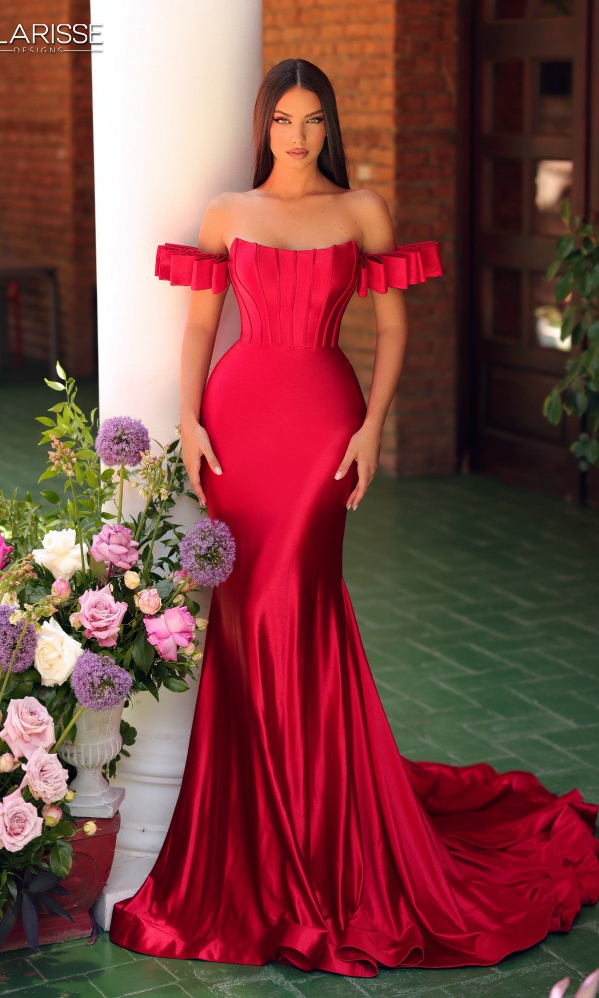 A woman stands indoors beside a floral arrangement, wearing the Long Prom Dress: Clarisse Designs 811160 in red; the fitted, off-the-shoulder gown is set against green flooring and brick walls.